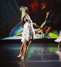 Mariana Tejeda dancing on stage in a white flowy dress with colorful shapes on screen behind her
