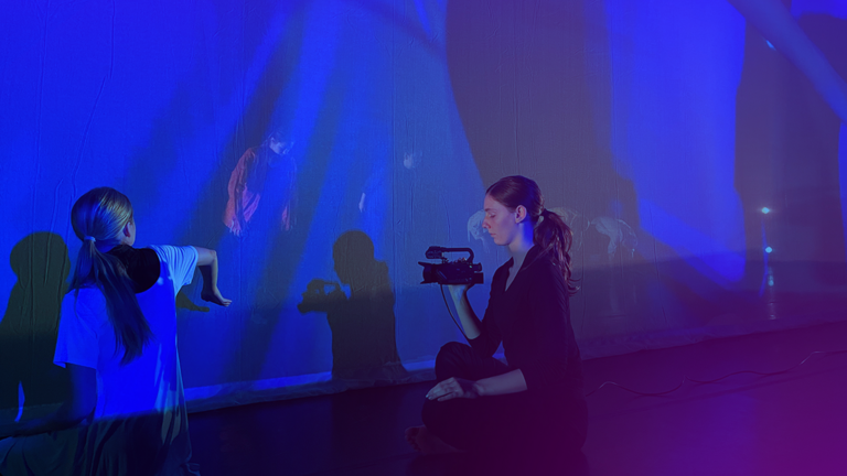 Dancer holding video camera and recording another dancer in performance.