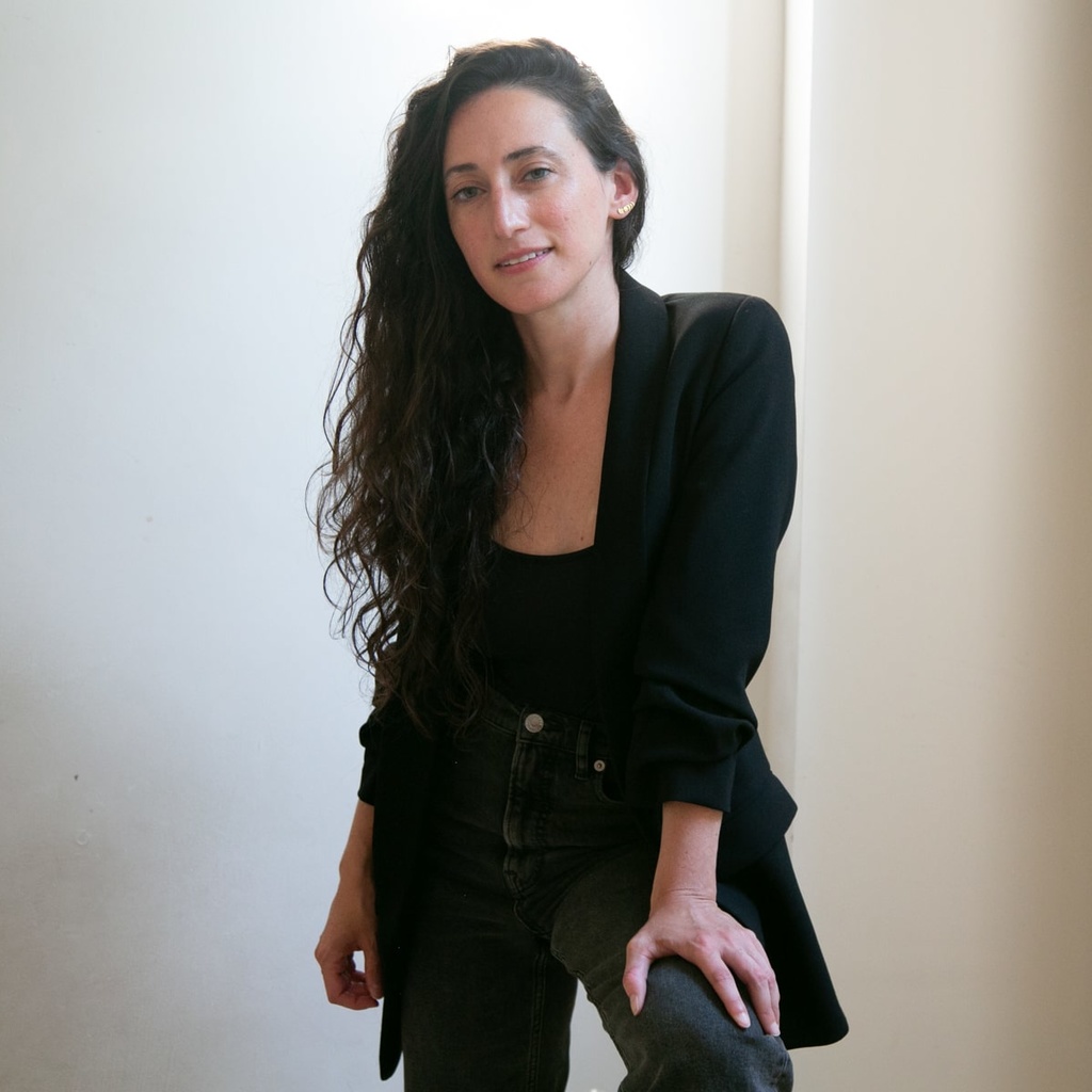 Photo of Danielle Russo from head to mid-thigh, facing the camera and smiling. Russo is leaning slightly forward, left hand resting on raised left knee. A white wall with a small white pipe running up the wall can be seen in the background.