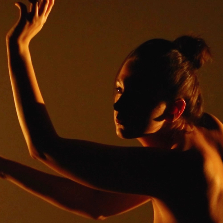 A close-up, action picture of Zoe Miller dancing in orange light with dramatic shadows. Zoe is facing away from the camera, looking off to the left.