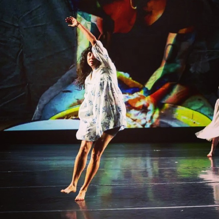 Mariana Tejeda dancing on stage in a white flowy dress with colorful shapes on screen behind her