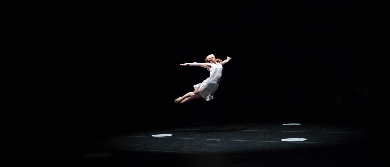 a light-skinned woman dancer in light-colored dress, suspended in the air agains a start black background