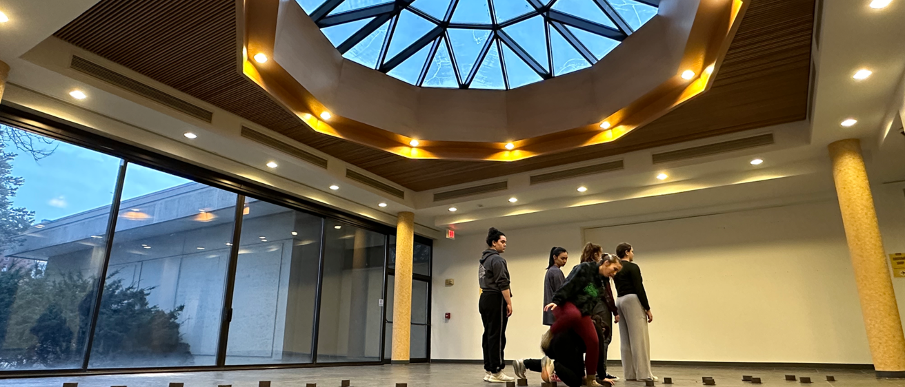 A photo of an open room with one glass wall and a geometric skylight. Small cubes are on the floor, forming a diagonal line across the room. Six dancers are next to each other in the middle of the room. Four dancers are forming a line facing a corner of the room. One dancer is crawling on the floor, while the other dancer is slightly bent over with one leg raised.