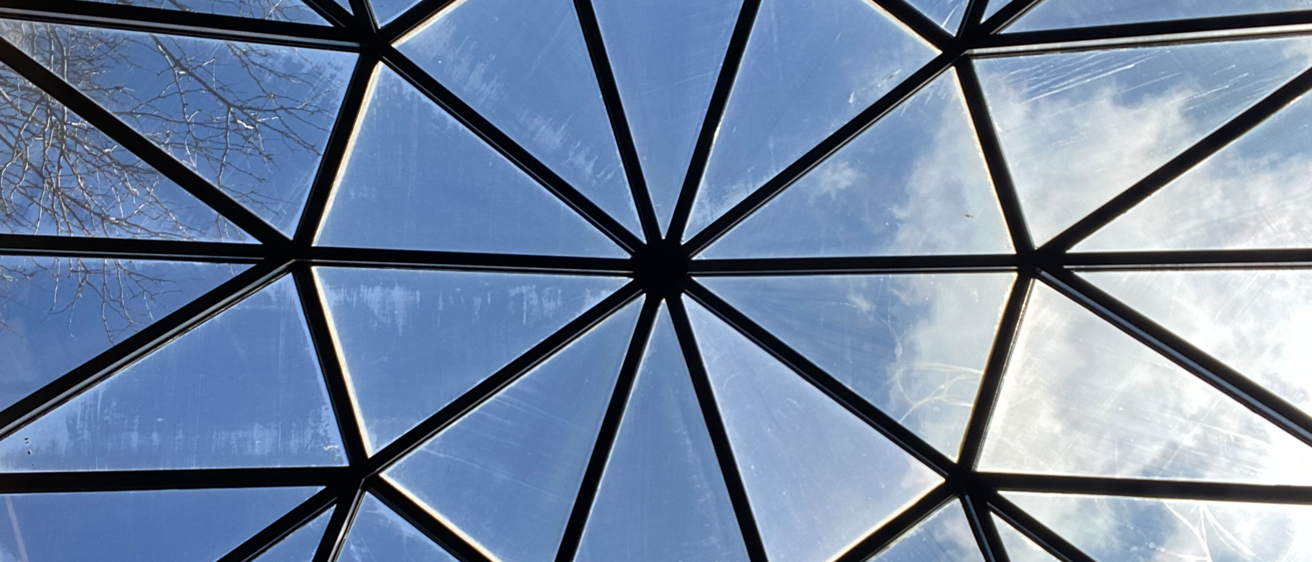 A photo from the perspective of someone looking up at a glass, geometric skylight. The skylight is a decagon divided up into triangles that meet in the middle. Visible through the skylight is a blue sky with clouds and tree branches peeking out at the bottom.