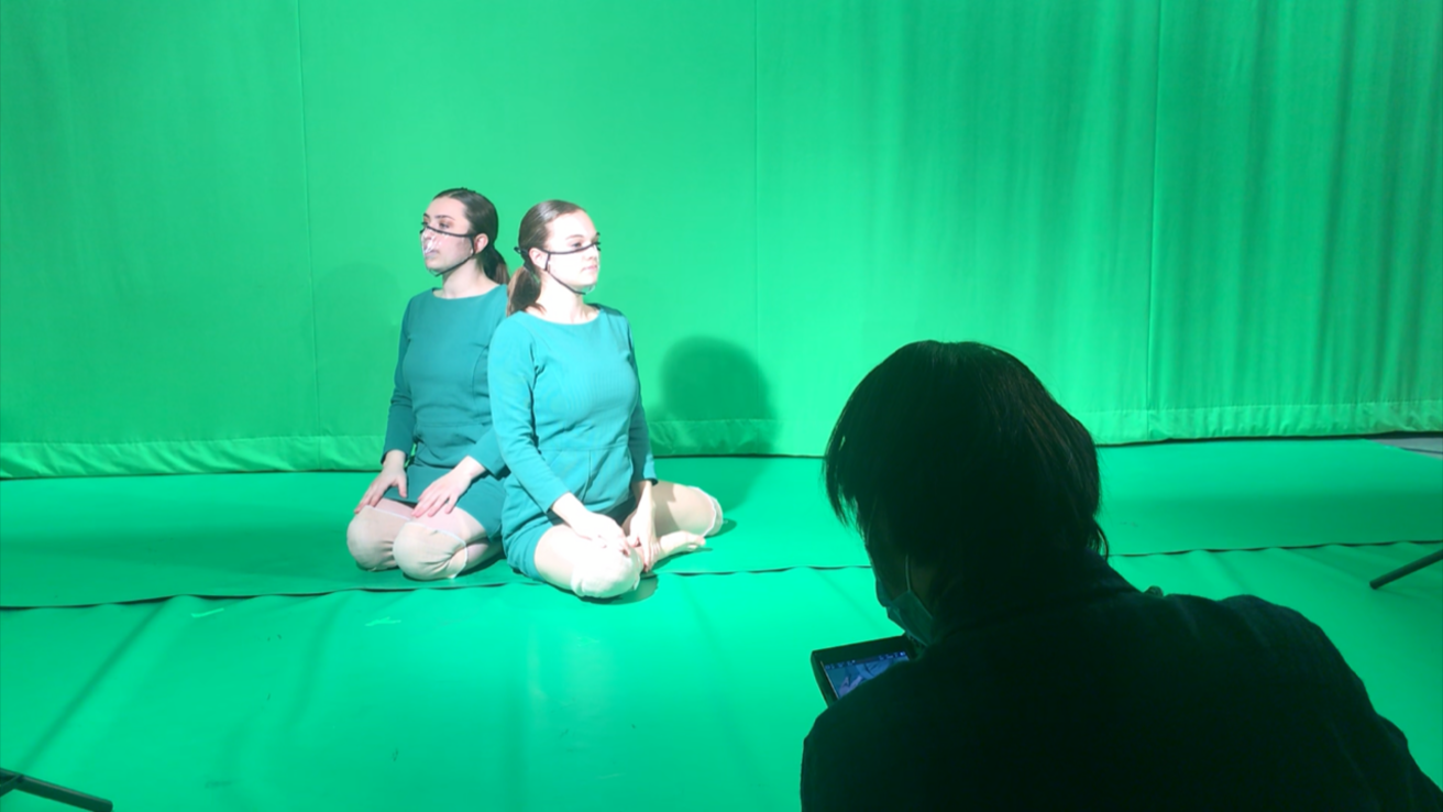 Two dancers kneeling in front of a green screen, back to back